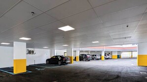 Un aparcamiento de varios niveles con varios coches estacionados, pilares de hormigón e iluminación colgante.
