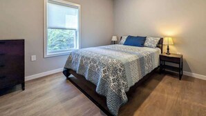 Un dormitorio con un ventanal grande, una cama con ropa de cama estampada, una mesita de noche con lámpara y un aparador.