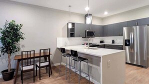 Una cocina moderna con una isla central, taburetes de bar, una mesa de comedor con sillas, una planta en maceta y electrodomésticos de acero inoxidable.