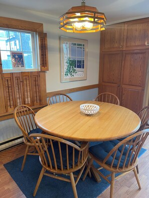 Een eetruimte met een ronde houten tafel, zes houten stoelen, een blauw vloerkleed, een houten kast en een raam met luiken.