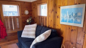 Une pièce chaleureuse avec un canapé bleu foncé, deux coussins blancs, un mur en bois, un tableau encadré, une horloge et un panier en osier.