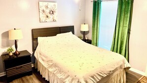 Un dormitorio con cama, dos mesitas de noche, ventilador de techo, ventana con cortinas verdes y una obra de arte colgada en la pared.