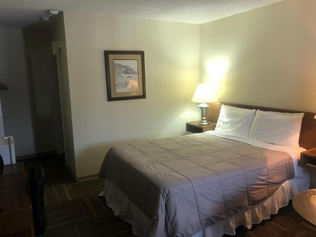 Una habitación de hotel con una cama, una mesita de noche, una lámpara, un cuadro en la pared y una puerta que da a otra habitación.