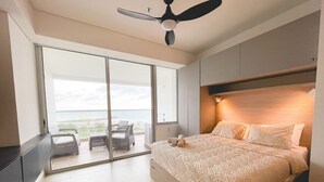 Un dormitorio moderno con una cama grande, un ventilador de techo y vistas al océano a través de puertas de vidrio del suelo al techo.