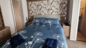 A bedroom with a bed, a headboard with a leaf pattern, and a wooden wardrobe. - Mini Lodge Robin Hood Holiday Park (Rhyl)