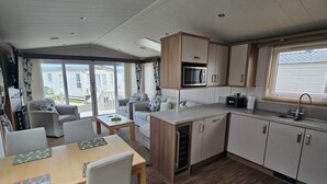A compact kitchen with a microwave, sink, and wooden cabinets. There is a dining area with a table and chairs, and a sliding glass door leading to a balcony. - Mini Lodge Robin Hood Holiday Park (Rhyl)