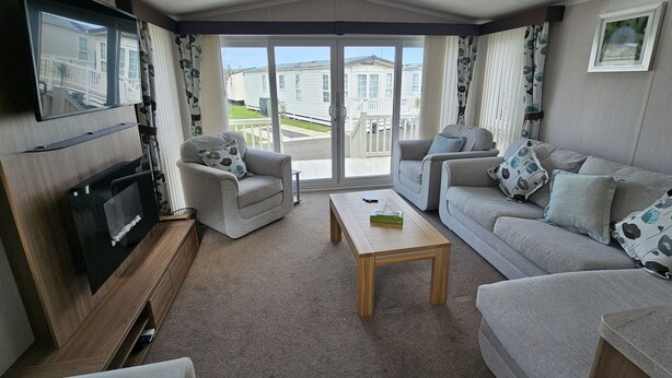 A living room with a TV, sofas, a coffee table, and a sliding glass door leading to a balcony. - Mini Lodge Robin Hood Holiday Park (Rhyl)