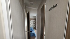 A narrow hallway with doors on both sides leading to a bedroom with a bed and a patterned headboard. - Mini Lodge Robin Hood Holiday Park (Rhyl)