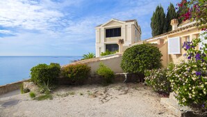 Un bâtiment situé sur la côte, avec un premier plan sablonneux, un mur de pierre et une vue sur la mer.