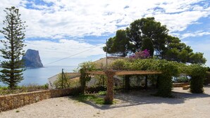 Une propriété située sur la côte, dotée d’un mur en pierre, d’une pergola couverte de plantes grimpantes et offrant une vue sur la mer et une île rocheuse.