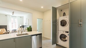 A modern kitchen with a dishwasher, a sink, and a built-in oven. There is a dining area with a table and chairs, and a view of the city through large windows. - HOMA-Comfort&Style-2BR-Spacious-Natural Light-FeelHome-A/C-SelfCheckIn-Metro (Montréal)
