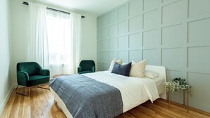 A modern bedroom with a large bed, two green chairs, a wooden floor, and a light-colored wall with paneling. - HOMA-Comfort&Style-2BR-Spacious-Natural Light-FeelHome-A/C-SelfCheckIn-Metro (Montréal)