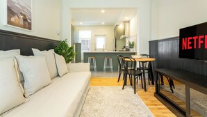 A modern living room with a sofa, a dining area with stools, a TV, and a kitchen in the background. - HOMA-Comfort&Style-2BR-Spacious-Natural Light-FeelHome-A/C-SelfCheckIn-Metro (Montréal)