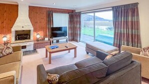 A living room with a fireplace, television, and large windows. - Cozy Highland Retreat With Stunning Views (Invergarry)