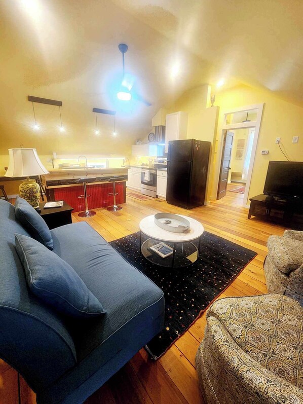 A living room with a sofa, a coffee table, a television, and a kitchen area in the background. - Cozy Loblolly Loft in charming Summerville (Summerville)
