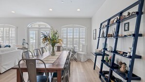 A dining area with a wooden table, chairs, and a vase of flowers. There are white walls and a bookshelf with various items.