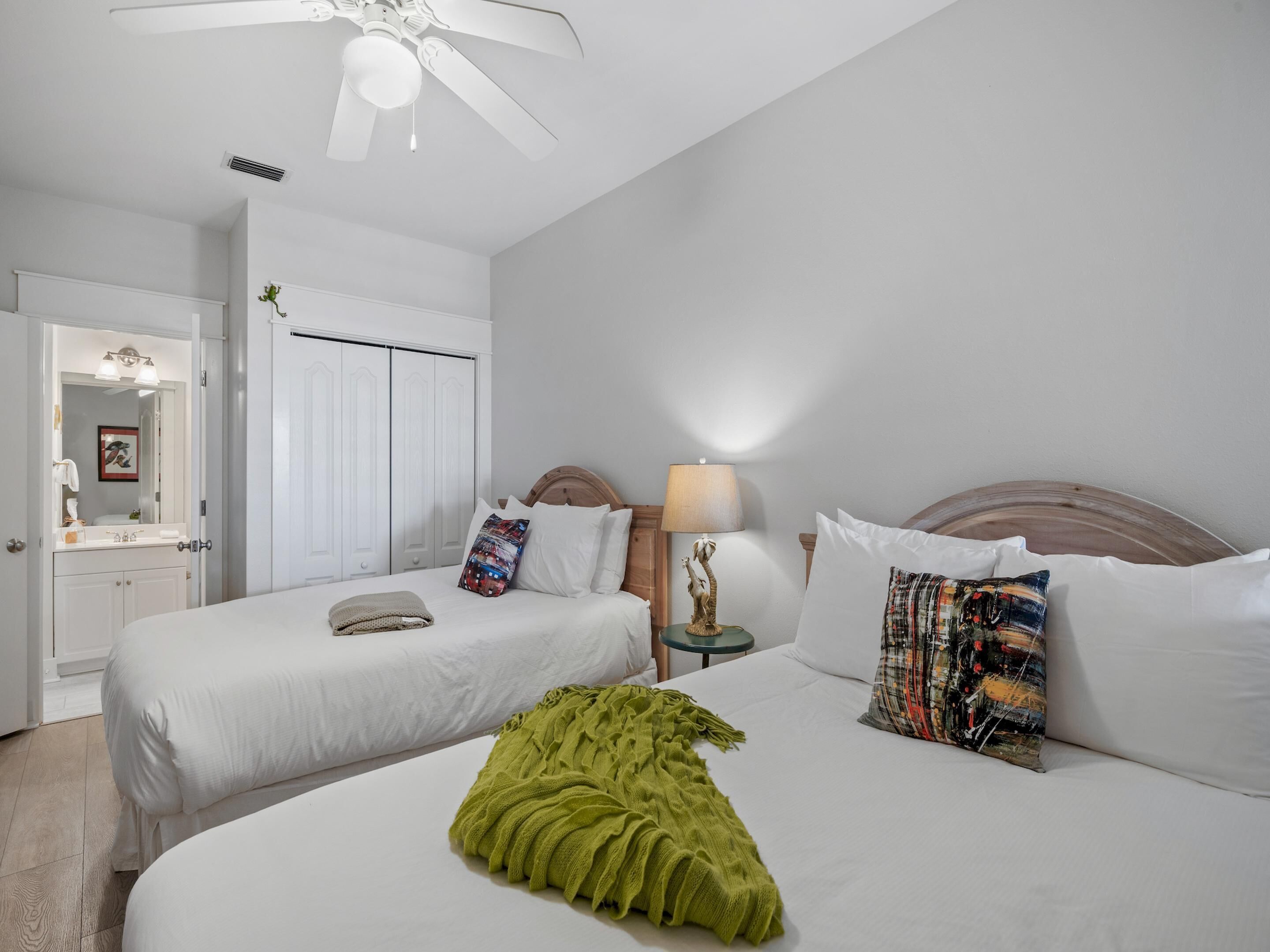 A hotel room with two beds, a ceiling fan, a nightstand with a lamp, and a bathroom with a mirror and sink.