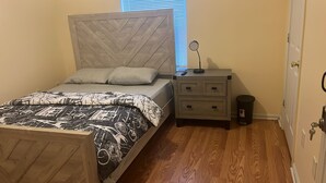 A bedroom with a bed, bedside tables, a trash bin, and a window with blinds.