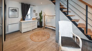 Una sala de estar moderna con piso de madera, un sillón de mimbre blanco, un aparador blanco, un tapete redondo tejido, una escalera con escalones de madera y una planta de cactus.