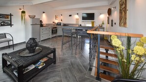 Una cocina moderna con una isla central, una zona de comedor con taburetes de bar, una mesa de centro negra con revistas y una escalera con barandilla de madera.