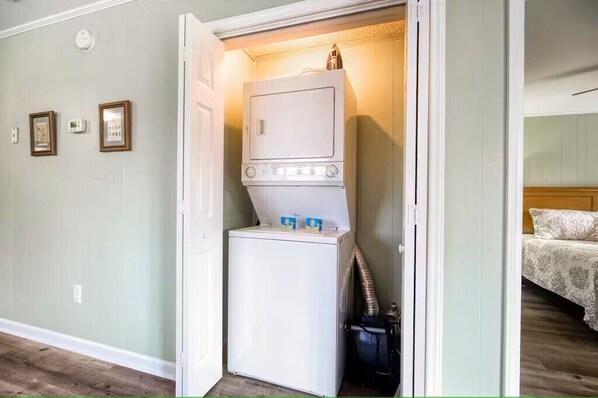 A laundry room with a stacked washer and dryer, a small trash can, and a door leading to a bedroom with a bed.