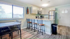 A modern kitchen with a dining area, featuring a large window, bar stools, and a trash can. - North Myrtle Retreat, 2bd 2ba apartment in 3unit building 1-block from the Beach (North Myrtle Beach)