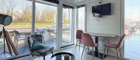 Ein modernes Wohnzimmer mit großen Fenstern, einem Flachbildfernseher, einem Essbereich mit rosa Stühlen und einem runden Couchtisch.