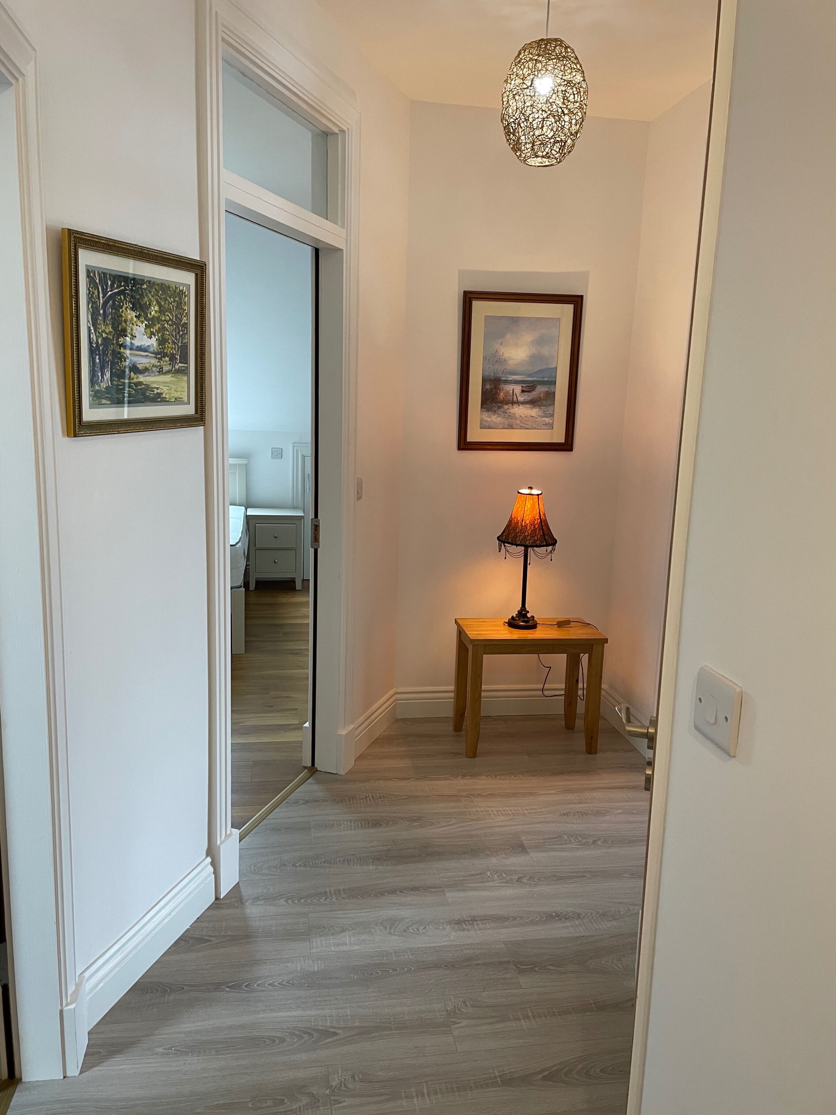 A hallway with wooden flooring, a table with a lamp, and two framed pictures on the walls.