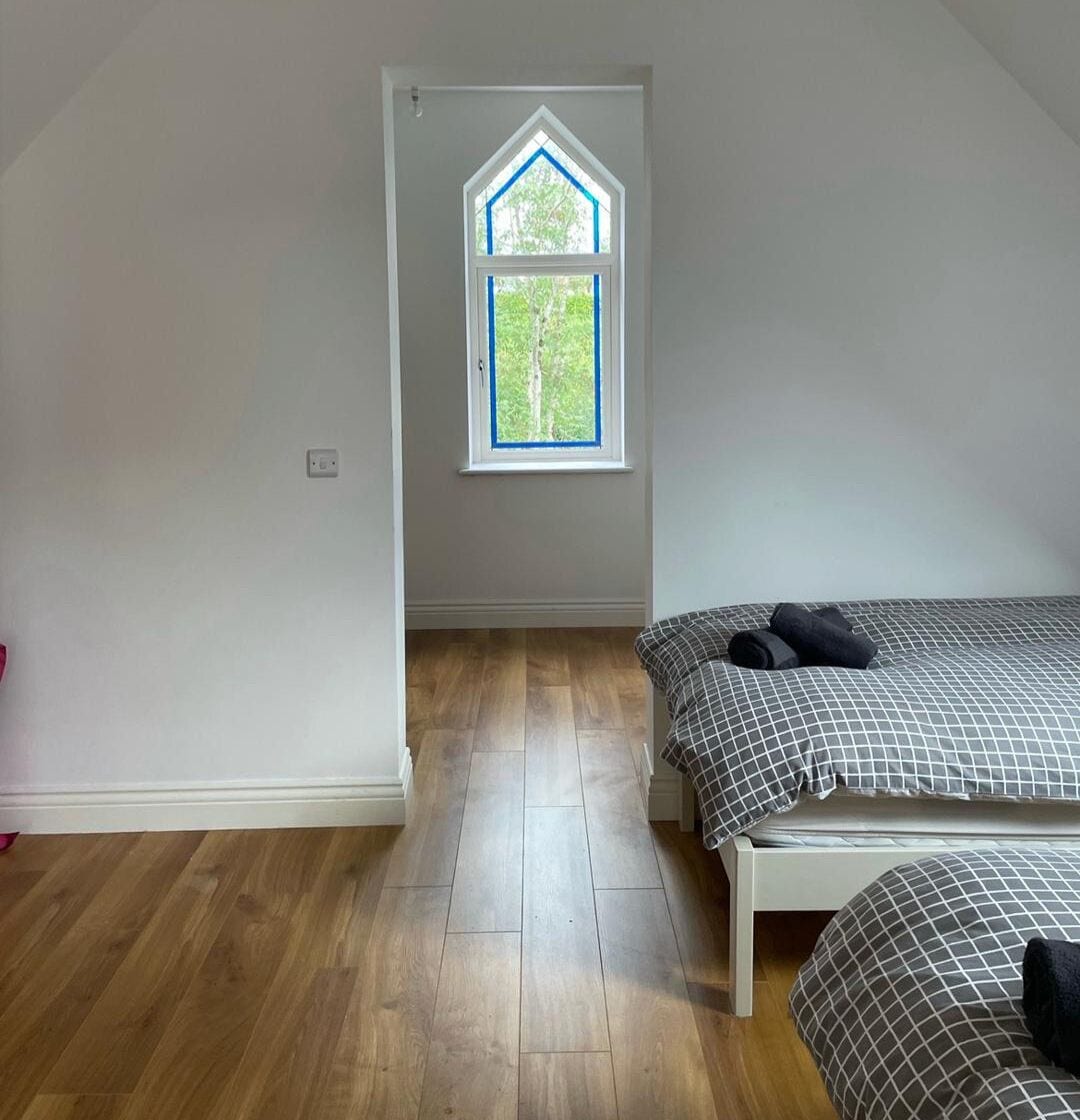A bedroom with a sloped ceiling, a window with blue trim, a wooden floor, and a bed with a checkered bedspread.