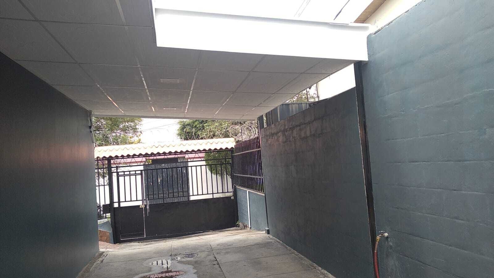 Un callejón estrecho con una entrada porticada, una valla metálica y un edificio con tejado de tejas al fondo.