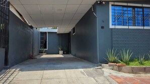 Une ruelle étroite avec un sol en béton, un bâtiment gris sur la droite et un mur bleu avec une fenêtre.