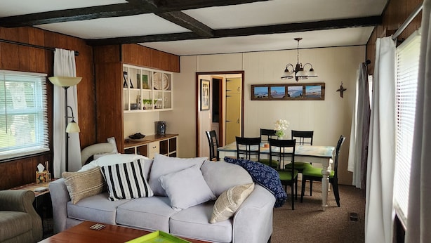 A cozy living room with a grey sofa, a wooden dining table with chairs, and a kitchen area with cabinets and a hanging light fixture.