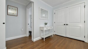 Un pasillo con piso de madera, paredes blancas, una consola blanca con un florero y un espejo.