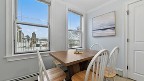Comedor con una mesa de madera y sillas blancas, ventanas amplias con vistas al exterior y un cuadro en la pared.