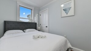 Una cama bien tendida con sábanas blancas y toallas enrolladas, un cabecero tapizado en gris, una ventana con vistas al cielo y una obra de arte abstracta enmarcada en la pared.