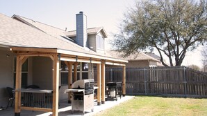 Une terrasse couverte avec un barbecue et un coin salon, une clôture en bois et un arbre en arrière-plan.