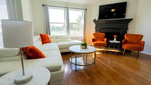 Un salon avec un canapé blanc, des fauteuils orange, une table basse ronde, un téléviseur fixé au-dessus d’une cheminée et de grandes fenêtres avec des rideaux.