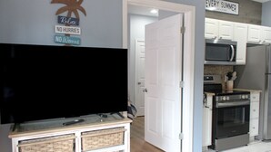 A living room with a flat-screen TV on a stand, a kitchen with white cabinets and stainless steel appliances, and a door leading to another room.
