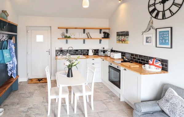 A modern kitchen with white cabinetry, a wooden countertop, and a dining area with white chairs. There is a clock on the wall and a shelf with various items. - Moments From Beach/lodge/nature Reserve/sandilands (Mablethorpe)