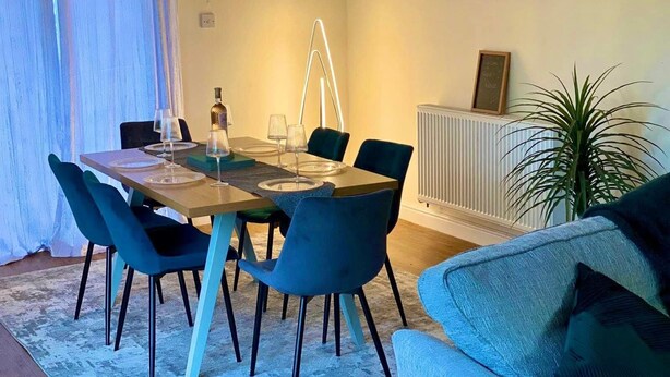 Zona pranzo con tavolo apparecchiato per quattro persone, sedie blu, termosifone e un quadro che raffigura un panorama cittadino.