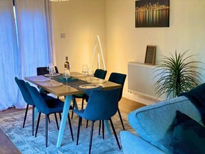 A dining area with a table set for four, blue chairs, a radiator, and a cityscape painting on the wall. - Radway Retreat - Cozy Countryside House (Warwick)