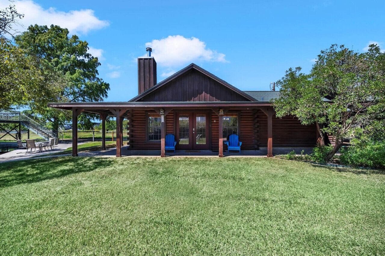 A log cabin with a porch, two blue Adirondack chairs, and a covered patio area.
