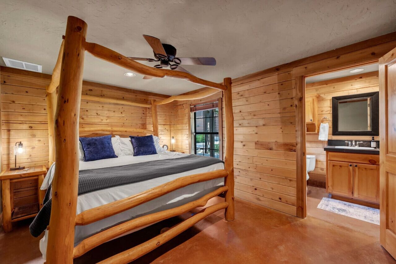 A log cabin-style bedroom with a four-poster bed, a ceiling fan, and a view of the outside through a window.