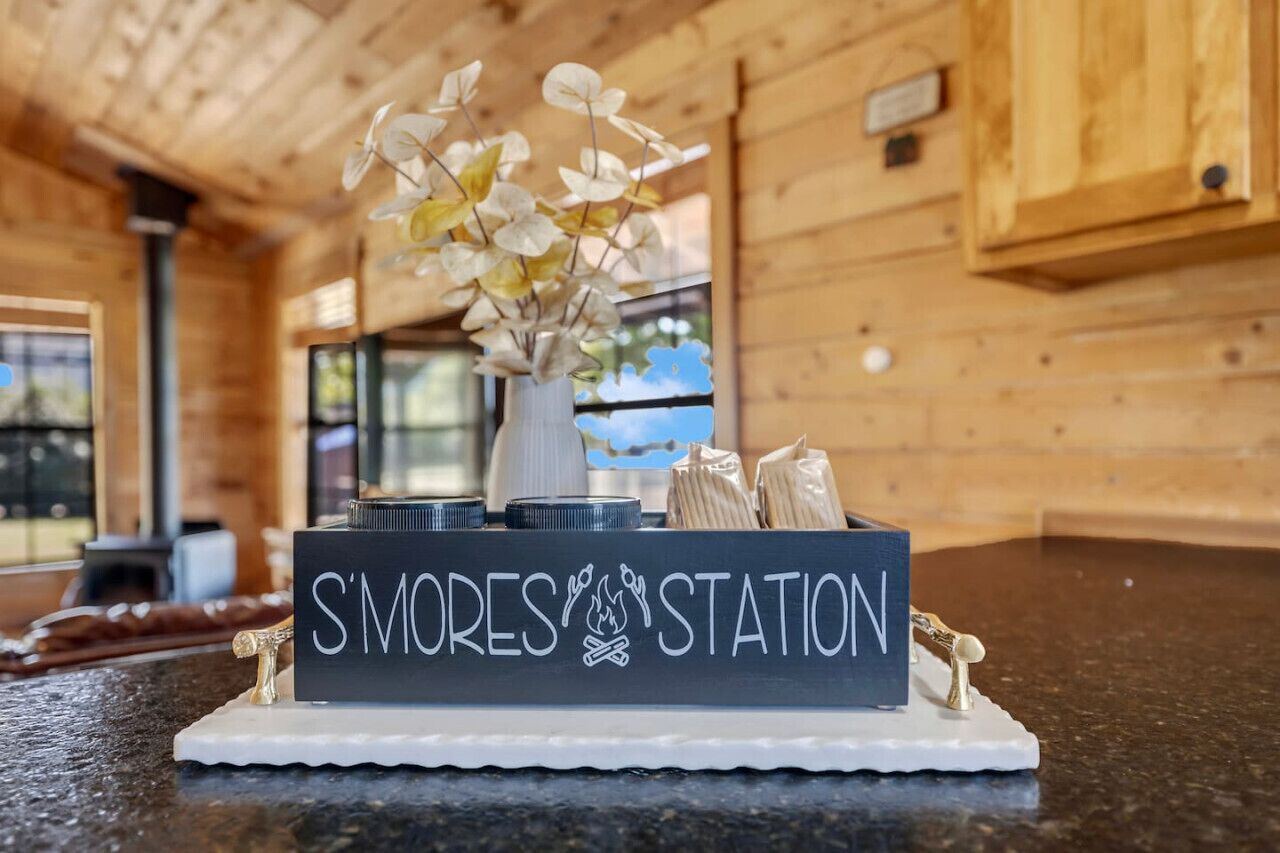 A wooden cabin interior with a sign reading "S'MORES STATION" on a table, surrounded by camping gear and a decorative plant.
