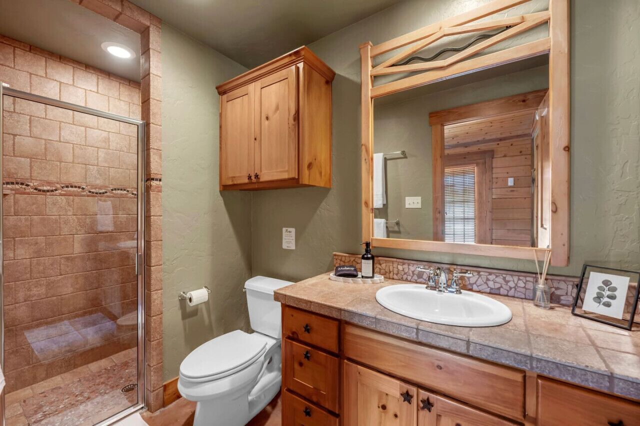 A bathroom with a glass-enclosed shower, a white toilet, wooden cabinets, a large mirror, and a sink with a stone countertop.