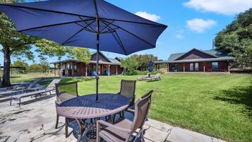 A patio with a round table and chairs, a large umbrella, and a wooden cabin in the background.