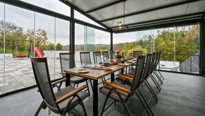 Un coin repas avec une table dressée pour six personnes, entourée de chaises à dossier en treillis, et de grandes fenêtres donnant sur un terrain de jeux et des arbres.