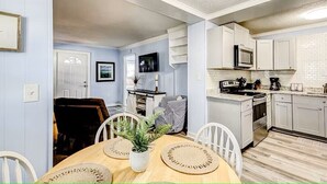 A modern kitchen with white cabinets, a microwave, and a stove. There is a dining area with a wooden table and chairs, and a potted plant on the table. - North Myrtle Getaway, 2bd 2ba bottom unit in 3unit building 1-block from beach (North Myrtle Beach)