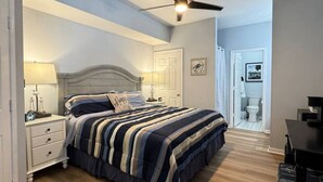 A bedroom with a bed, nightstands, a ceiling fan, and a door leading to another room. - The Suite at 125 Royal Postes Rd (Sunset Beach)
