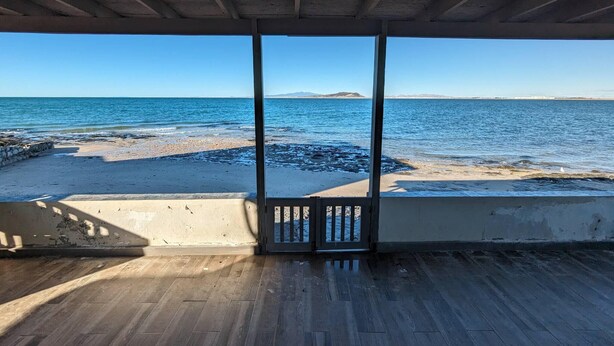 A room with a wooden floor, a concrete wall, and a view of the sea through a glass door.